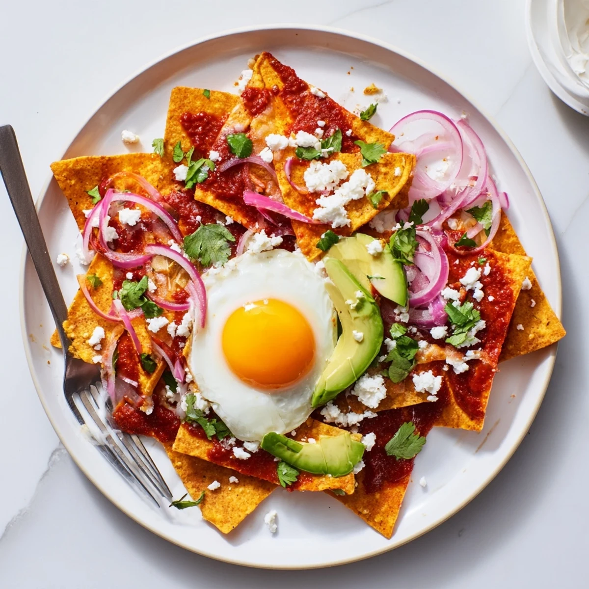 Crispy fried corn tortilla chips coated in zesty salsa verde, topped with a runny yolk egg, crumbled queso fresco, sliced avocado, and fresh cilantro garnish for a vibrant Mexican breakfast dish.