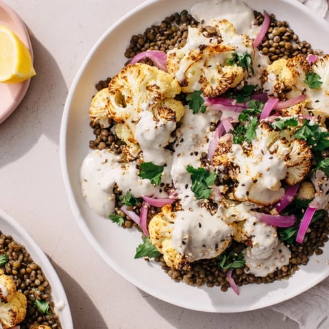 Golden roasted cauliflower florets with caramelized edges rest atop a vibrant lemon lentil salad flecked with red onion and parsley, all drizzled with creamy cumin tahini yogurt.