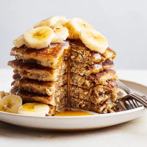 Stack of warm, golden-brown banana oat pancakes topped with fresh berries and a drizzle of maple syrup on a rustic plate.